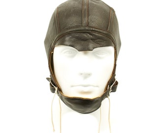 May include: A brown leather aviator helmet with a chin strap and tie strings. The helmet has a simple design with no embellishments.
