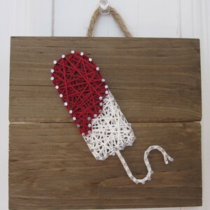 May include: A red and white popsicle design created with string art on a wooden board. The popsicle is wrapped in white string with a red string top and a white string drip.