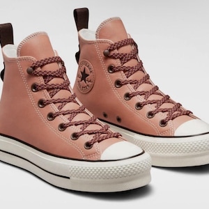 May include: A pair of high-top sneakers in a dusty rose color. The shoes feature a white platform sole, white toe cap, and brown laces. The iconic Converse star logo is visible on the side. The back of the shoe has a brown pull tab.
