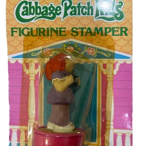 May include: Vintage Cabbage Patch Kids figurine stamper in original packaging. The packaging is yellow, pink, and blue, with the words "Cabbage Patch Kids" and "Figurine Stamper" printed on the front. The stamper features a red-haired doll.