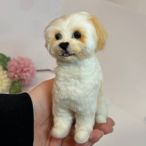 May include: A white and tan needle felted dog figurine with black eyes and a pink tongue. The dog is sitting on a person's hand.
