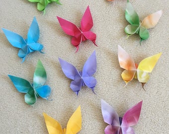 May include: Nine colourful origami butterflies are displayed. The butterflies are in shades of green, blue, pink, purple, and yellow. The paper butterflies are arranged on a neutral surface, showing their folded wings and delicate antennae.