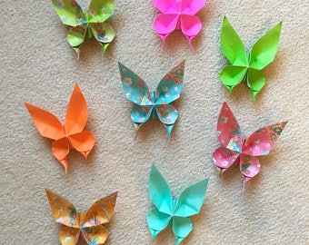 May include: Eight origami butterflies in various colors and patterns, including green, pink, orange, blue, and yellow, are displayed on a white fuzzy surface.
