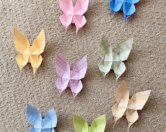 May include: Eight origami butterflies in various colors and patterns, including pink, yellow, blue, and green, are displayed on a beige textured surface.