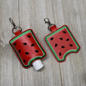 May include: Two red and green watermelon-patterned hand sanitizer holders with silver keychains. The holders are made of fabric and have a stitched design. One holder has a white hand sanitizer bottle inside.