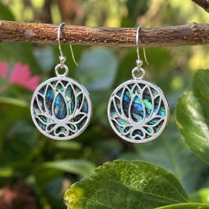 May include: A pair of silver-toned drop earrings featuring a circular frame with a cut-out lotus flower design. The centre of each earring is inlaid with iridescent abalone shell displaying vibrant blue and green hues, suspended from silver hooks.
