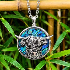 A silver-toned pendant necklace featuring a Highland cow design. The pendant has a circular frame with a blue and green iridescent background. The cow's head is detailed in silver, with horns extending outward. The necklace chain is also silver.