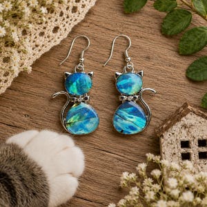May include: A pair of silver-toned drop earrings shaped like cats, featuring iridescent blue and green inlay material. Each earring has a hook-style closure and a small clear stone accent at the neck of the cat figure, with a smooth, polished finish.