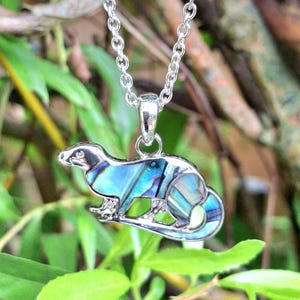 Puede incluir: Un collar plateado con un colgante con forma de peque&ntilde;o animal, posiblemente un hur&oacute;n o una comadreja. El colgante est&aacute; incrustado con concha de abul&oacute;n iridiscente azul y verde. La cadena es de estilo eslabones delicados, sobre un fondo de follaje verde borroso.