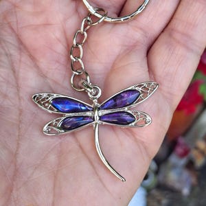 Può includere: Un portachiavi in metallo color argento a forma di libellula, caratterizzato da quattro ali con inserti iridescenti viola e blu. La libellula è collegata a un anello circolare standard tramite una corta catenella in metallo.