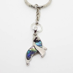 Può includere: Un portachiavi color argento con un ciondolo a forma di foca. Il ciondolo a forma di foca ha una finitura argentata lucida e presenta intarsi iridescenti di conchiglia di abalone, che mostrano sfumature blu, verdi e viola. Il portachiavi include un anello diviso e una catena.
