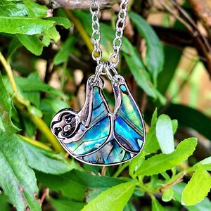 Abalone sloth necklace hanging from a tree branch outdoors, featuring an iridescent blue and green paua shell sloth pendant on a silver trace chain with a lake and woodland backdrop.