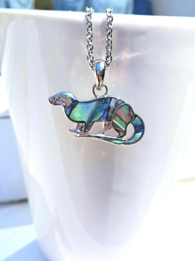 Otter necklace with colourful abalone shell pendant displayed on a white ceramic mug, shown on a silver-tone trace chain.