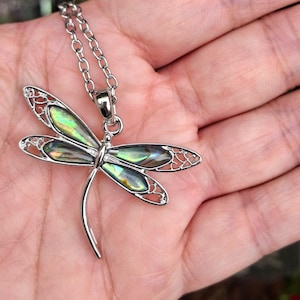 Puede incluir: Un collar con colgante de lib&eacute;lula plateada. La lib&eacute;lula tiene alas iridiscentes verdes y azules y un dise&ntilde;o calado delicado. La cadena del collar tambi&eacute;n es plateada.