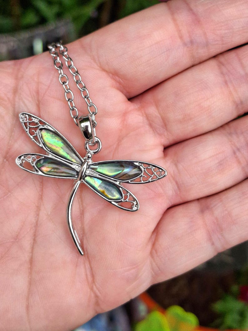 Puede incluir: Un collar con colgante de lib&eacute;lula plateada. La lib&eacute;lula tiene alas iridiscentes verdes y azules y un dise&ntilde;o calado delicado. La cadena del collar tambi&eacute;n es plateada.