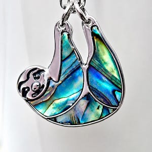 Abalone sloth necklace hanging from a tree branch in a garden setting, featuring an iridescent paua shell sloth pendant on a silver trace chain with green foliage and natural daylight.