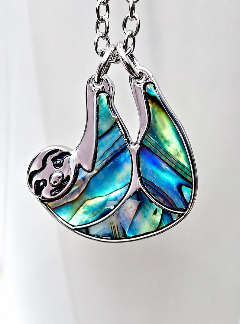 Abalone sloth necklace hanging from a tree branch in a garden setting, featuring an iridescent paua shell sloth pendant on a silver trace chain with green foliage and natural daylight.