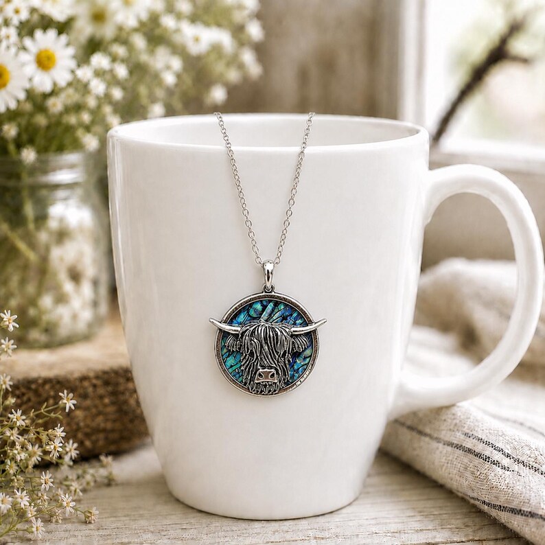 Abalone Highland Cow necklace with trace chain displayed on a white mug in a soft rustic kitchen scene with daisies and neutral decor.