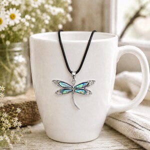 Abalone shell dragonfly pendant necklace on a black cord displayed on a white mug in a rustic floral setting.