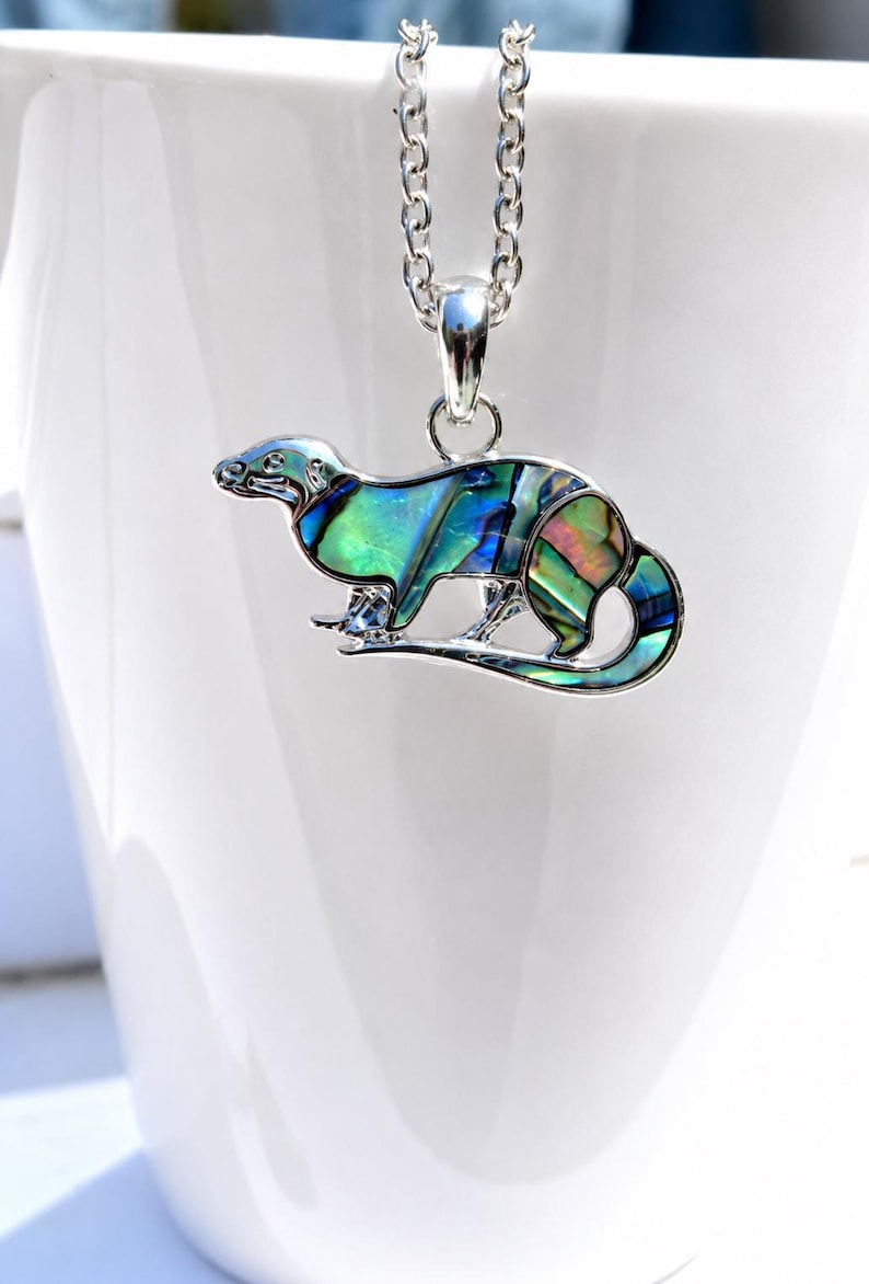 Otter necklace with colourful abalone shell pendant displayed on a white ceramic mug, shown on a silver-tone trace chain.