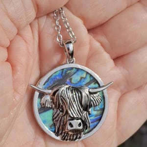 K&ouml;nnte beinhalten: Halskette mit silbernem Anh&auml;nger, der eine Highland-Kuh darstellt. Der Anh&auml;nger zeigt einen detaillierten Kuhkopf mit H&ouml;rnern, vor einem farbenfrohen Abalone-Muschel-Hintergrund. Die Halskette hat eine zarte Kette.