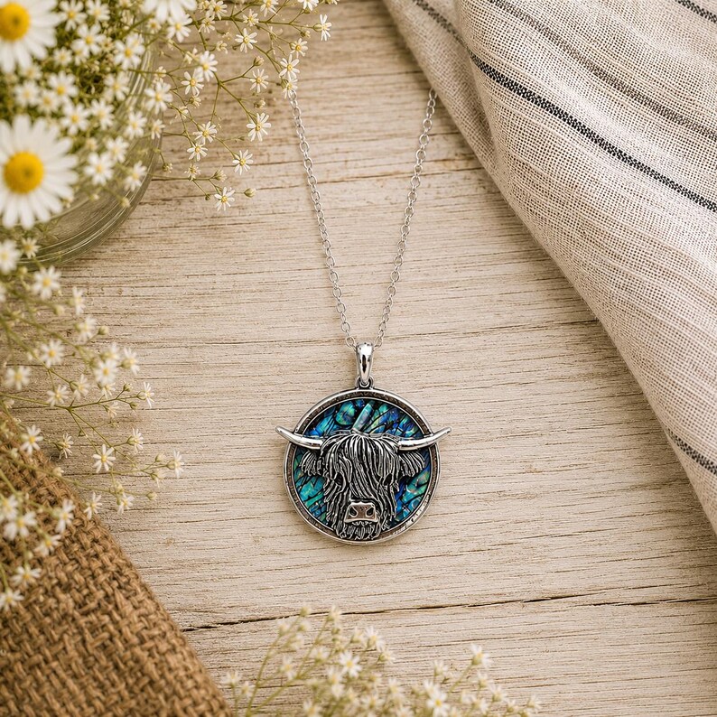 Abalone Highland Cow necklace with trace chain flat lay on a rustic wooden surface with wildflowers and soft neutral linen styling.