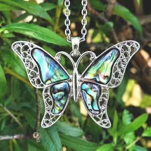 May include: A silver-toned butterfly pendant necklace featuring iridescent abalone shell inlays within the wings. The butterfly has intricate filigree metalwork detailing along the outer edges of the wings and hangs from a matching silver-toned chain.