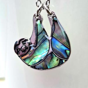 Abalone sloth necklace draped over a white ceramic mug outdoors, showcasing an iridescent blue and green paua shell sloth pendant on a silver trace chain with a natural garden backdrop.