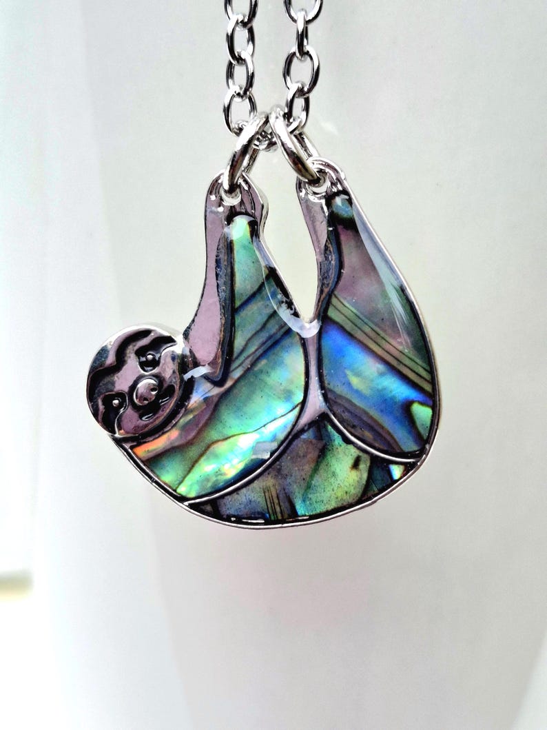 Abalone sloth necklace draped over a white ceramic mug outdoors, showcasing an iridescent blue and green paua shell sloth pendant on a silver trace chain with a natural garden backdrop.