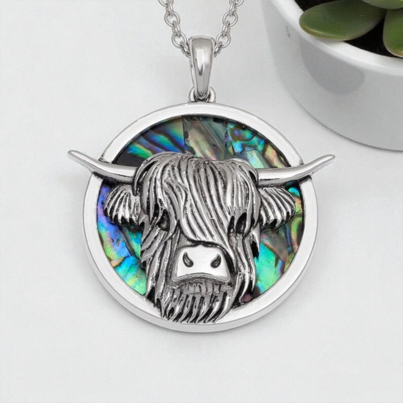 K&ouml;nnte beinhalten: Silberfarbene Anh&auml;nger Halskette mit einem Highland-Kuh-Design. Der Kopf der Kuh ist mit strukturiertem Fell und H&ouml;rnern versehen, vor einem Hintergrund aus schillerndem Abalone-Muschel. Der Anh&auml;nger h&auml;ngt an einer zarten Kette.