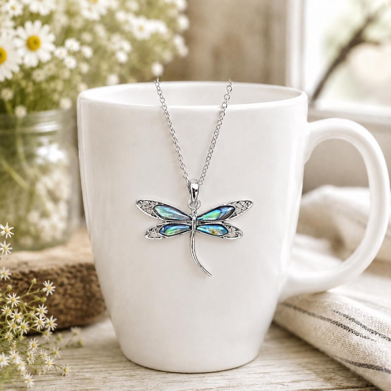 Abalone shell dragonfly necklace on a silver trace chain displayed on a white mug in a bright cottage-style setting with daisies.