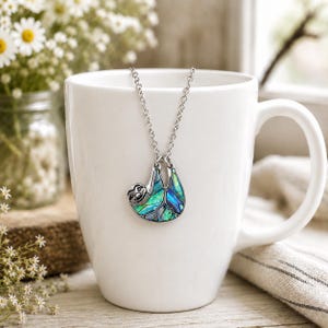 Abalone sloth necklace displayed on a white ceramic mug, featuring an iridescent blue and green paua shell sloth pendant on a silver trace chain with soft floral décor and natural lighting.