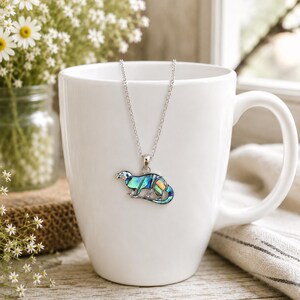 Otter necklace with colourful abalone shell pendant displayed on a white ceramic mug, shown on a silver-tone trace chain.