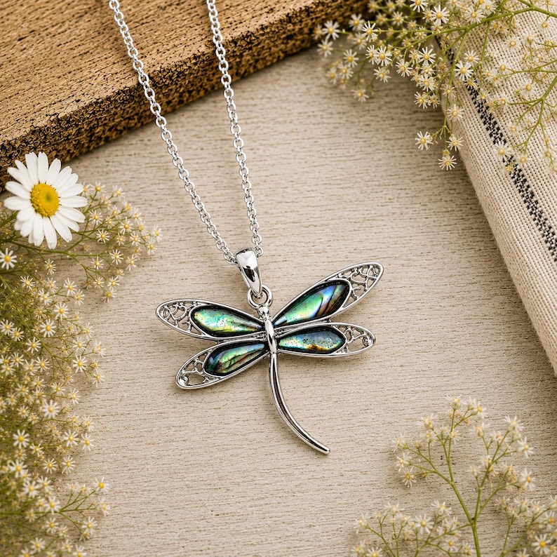 Silver dragonfly necklace with abalone shell inlay displayed on a rustic wooden surface surrounded by delicate white flowers.