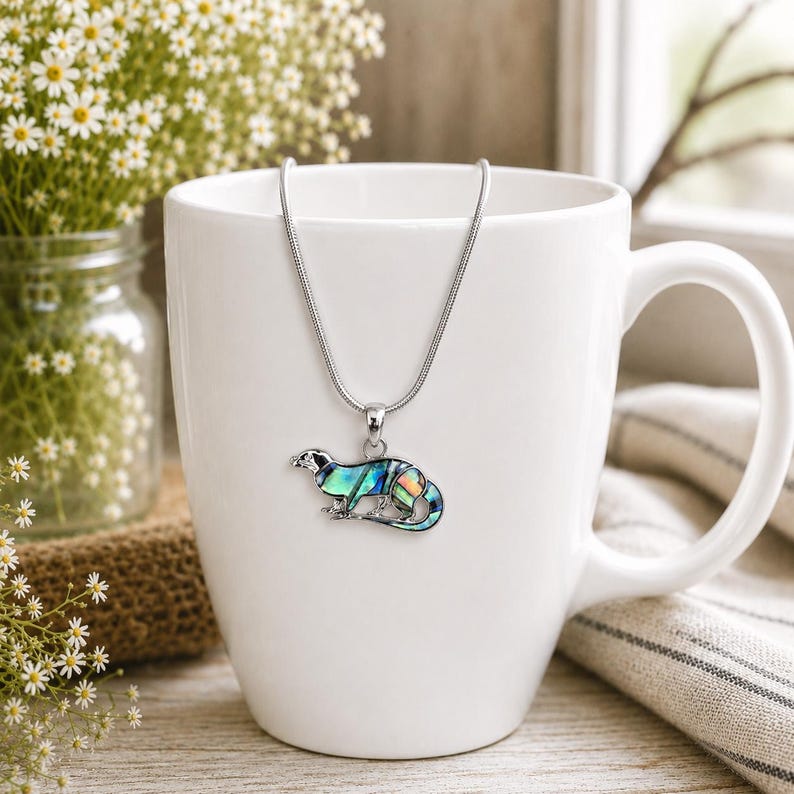 Abalone shell otter necklace displayed on a white mug with a silver-tone snake chain, showing vibrant blue and green paua shell detail.