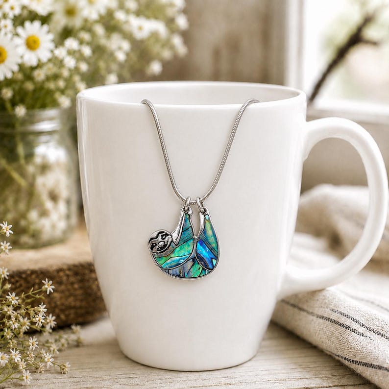 Abalone sloth necklace displayed on a white ceramic mug with a silver snake chain, featuring an iridescent blue and green paua shell sloth pendant in a soft floral setting.