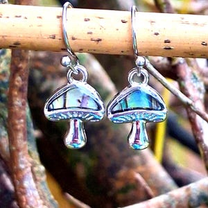 Könnte beinhalten: Ein Paar pilzförmige Ohrringe mit schillernden Kappen und silberfarbenen Stielen und Haken. Die Ohrringe hängen an einem Ast, der ihr einzigartiges Design und ihre Farbvariationen zeigt. Diese Ohrringe sind ein skurriles Accessoire.