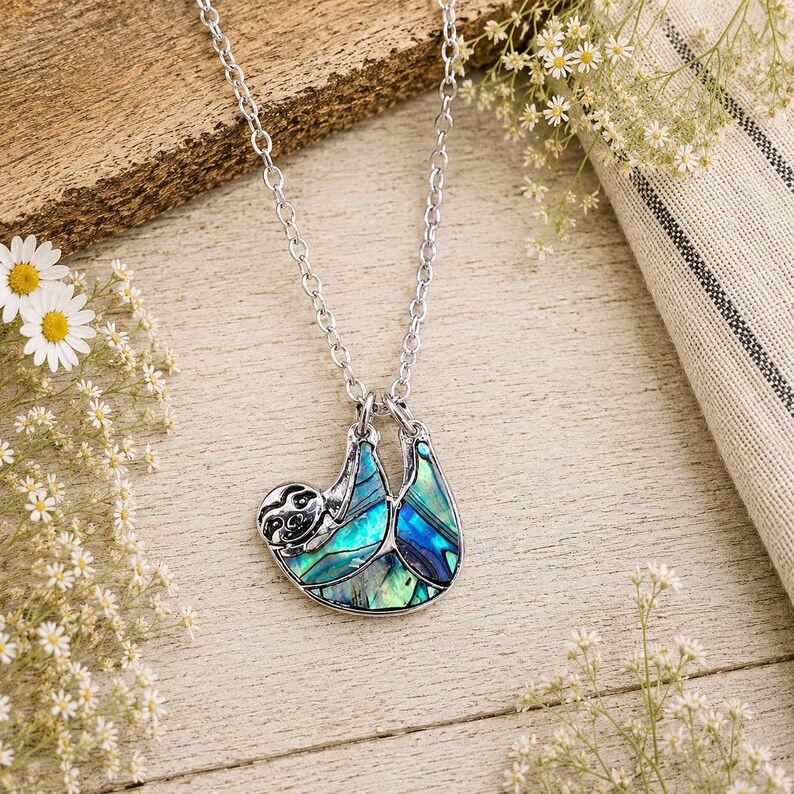 Abalone sloth necklace arranged on a rustic wooden surface with white flowers and neutral fabric accents, featuring an iridescent paua shell sloth pendant on a silver trace chain.
