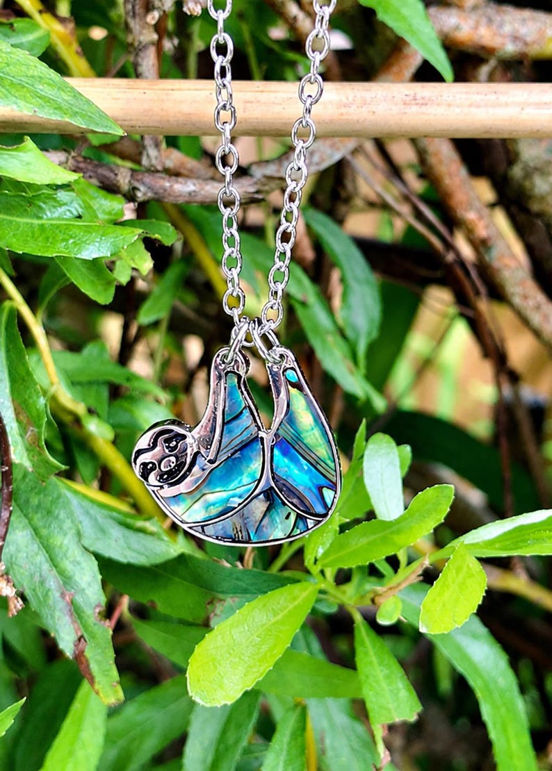Abalone sloth necklace hanging from a tree branch outdoors, featuring an iridescent blue and green paua shell sloth pendant on a silver trace chain with a lake and woodland backdrop.