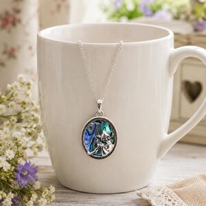 May include: A silver-toned pendant necklace featuring an oval-shaped inlay of iridescent abalone shell. The center of the pendant displays a detailed silver relief of a small creature holding a staff, suspended from a delicate silver chain.