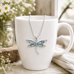 Abalone shell dragonfly pendant on a silver snake chain displayed on a white mug with soft floral decor and natural light.