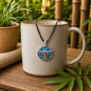 Highland cow abalone shell necklace displayed on a ceramic mug, featuring a silver-tone cow pendant with colourful paua shell inlay on a black cord necklace.