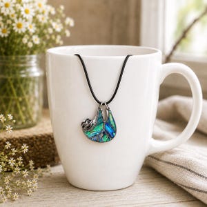 Abalone sloth necklace displayed on a white ceramic mug with a black cord necklace, featuring an iridescent blue and green paua shell sloth pendant with daisy floral décor.