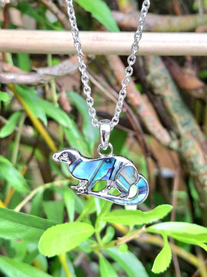 Puede incluir: Un collar plateado con un colgante con forma de peque&ntilde;o animal, posiblemente un hur&oacute;n o una comadreja. El colgante est&aacute; incrustado con concha de abul&oacute;n iridiscente azul y verde. La cadena es de estilo eslabones delicados, sobre un fondo de follaje verde borroso.
