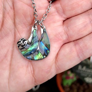 May include: A silver-toned metal pendant shaped like a sloth, featuring iridescent abalone shell inlay with vibrant green, blue, and purple hues. The sloth pendant hangs from a matching silver-toned metal link chain, showcasing a detailed face.