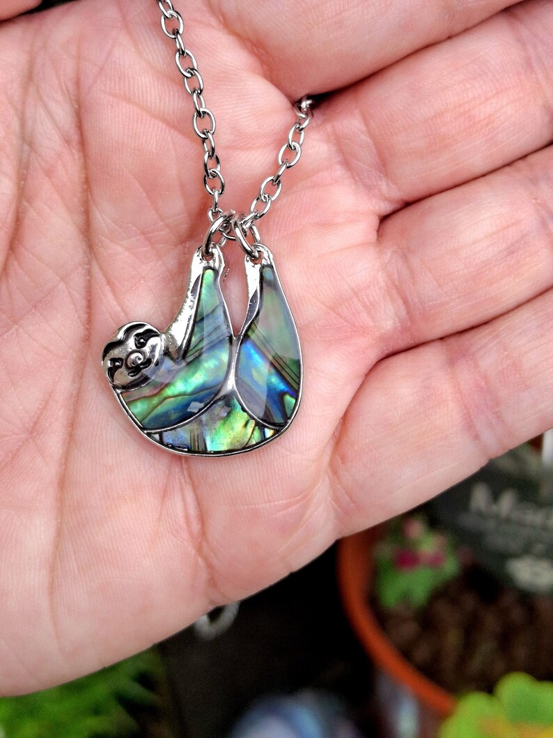 May include: A silver-toned metal pendant shaped like a sloth, featuring iridescent abalone shell inlay with vibrant green, blue, and purple hues. The sloth pendant hangs from a matching silver-toned metal link chain, showcasing a detailed face.