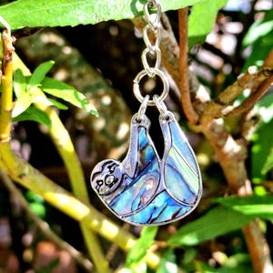 Può includere: Un ciondolo a forma di bradipo argentato con intarsi iridescenti di conchiglia blu e verde è appeso a una catena d'argento. Il ciondolo a forma di bradipo è sospeso su uno sfondo di foglie e rami verdi.