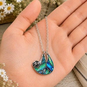 Abalone sloth necklace resting in an open hand, featuring a blue and green paua shell sloth pendant on a silver trace chain with a blurred daisy background and warm indoor styling.