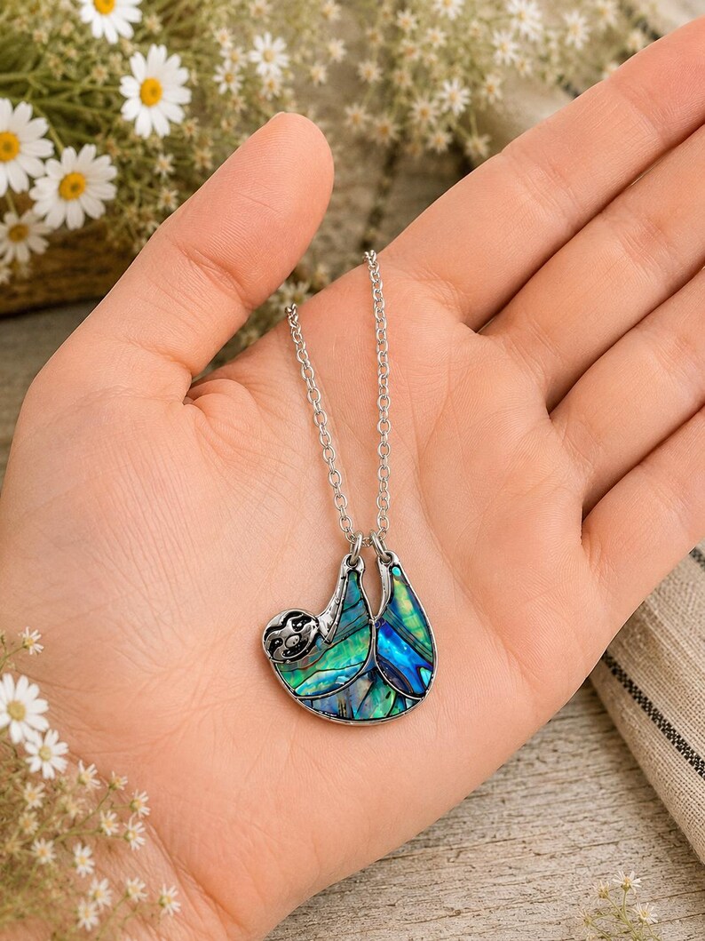 Abalone sloth necklace resting in an open hand, featuring a blue and green paua shell sloth pendant on a silver trace chain with a blurred daisy background and warm indoor styling.
