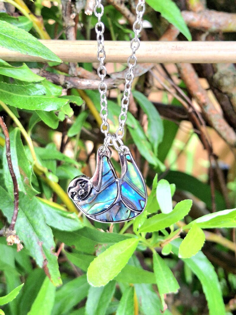 May include: A silver-toned metal pendant shaped like a sloth, featuring iridescent abalone shell inlay with blue and green hues. The sloth pendant hangs from a matching silver-toned metal link chain, showcasing a detailed face and limbs.